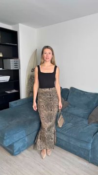 Schwarzes ärmelloses Oberteil und einen langen, taillierten Rock mit braunen Streifen, der am Saum leicht ausgestellt ist. Das Model hält eine kleine Handtasche und trägt beige Schuhe.