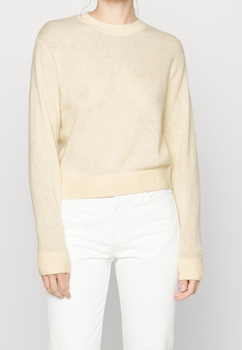 Pull crème à col rond, à manches longues et avec des bords côtelés. Fabriqué en tissu doux et texturé. Associé à un pantalon blanc.