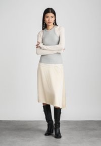 Canotta grigia a coste indossata sotto una maglia beige a maniche lunghe, abbinata a una gonna asimmetrica di raso crema e stivali neri al ginocchio.