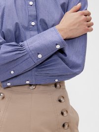 Camicia a righe blu e bianche con una vestibilità su misura, caratterizzata da bottoni e polsini bianchi, abbinata a pantaloni beige a vita alta con grandi bottoni.