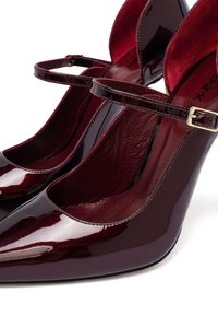 Tacones de charol burdeos con punta afilada, tira para el tobillo y un acabado suave. El interior presenta una textura suave.