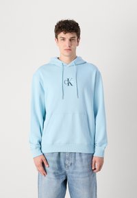 Sudadera con capucha azul claro, con un bolsillo tipo canguro en la parte frontal y puños acanalados, que presenta un pequeño logo "CK" en el centro.