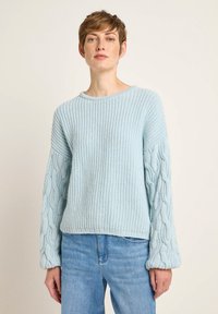 Hellblauer gestrickter Pullover mit geripptem Körper und Kabelstrickärmeln, ausgestattet mit einem runden Halsausschnitt und lockerer Passform. Kombiniert mit Jeans.