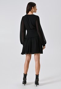 Zwarte jurk met doorschijnende lange mouwen, aansluitende taille en een uitlopende rok. Draagt het met zwarte hakken enkelschoenen met decoratieve studs.