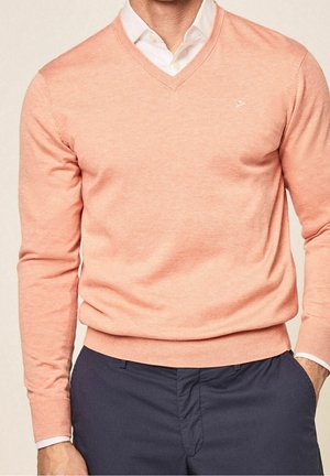 Uomo che indossa un maglione color pesca a scollo a V sopra una camicia bianca con colletto e pantaloni blu navy, con la mano destra in tasca.