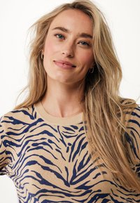 Beige tröja med marinblå zebra-ränder, rundad halsringning och mjuk textur. Har långt, vågigt hår och subtila hoop-örhängen.