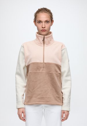 Un mannequin se tient devant un fond uni, vêtu d’un pull polaire à blocs de couleurs, avec un col montant zippé et un pantalon blanc.