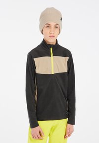 Fleece-Pullover in Schwarz und Taupe, mit einem halben Reißverschluss vorne, hohem Kragen und neon-gelbem Reißverschlussdetail. Kombiniert mit einer beigen Strickmütze.