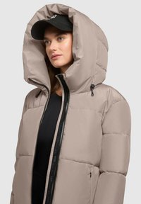 Bēšā pūkainā jaka ar lielu kapuci, melnu rāvējslēdzēju un sānu kabatām. Gluda tekstūra ar modernu dizainu, uzvilkta virs melna topa.