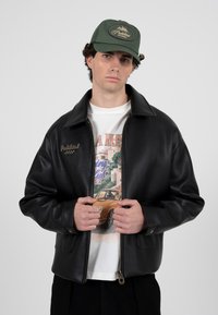 Mladý muž s zelenou čepicí, černou koženou bundou a bílým tričkem s potiskem ukazuje na veteránské auto a budovy.