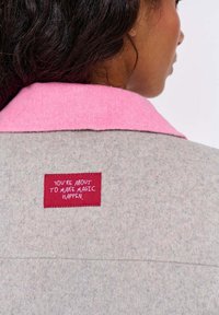 Šedý vlněný kabát s růžovým filcovým límcem. Na zadní straně se nachází červená textilní nášivka s bílým textem, který zní: "PRÁVĚ SE CHYSTÁTE UDĚLAT MAGII."