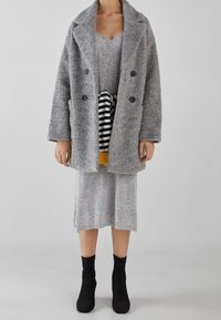 Grå ull dubbelknäppt kappa över en grå stickad klänning med en randig skjorta som tittar fram, ihop med rippade svarta strumpor.