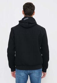 Uomo che indossa una giacca nera con cappuccio e etichetta del marchio sul cappuccio, visto di spalle, abbinata a jeans blu su sfondo semplice.