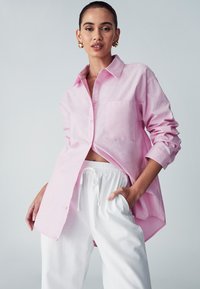 Camicia button-up rosa chiaro con taschino sul petto, maniche arrotolate e vestibilità rilassata, abbinata a pantaloni bianchi con vita regolabile.