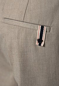 Jasnobeżowa tkanina o teksturowanej powierzchni; posiada małą kieszonkę z czarno-białą pętelką w paski oraz czerwonym przeszyciem.