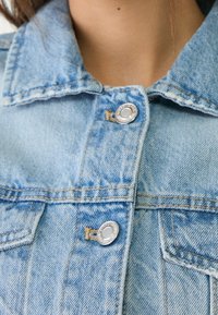 Ljusblå denimjacka med klassisk krage, silvriga tryckknappar och kontrasterande stygn; har en ficka på vänster sida.