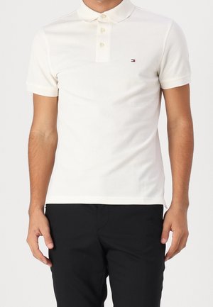 Homme portant un polo blanc uni à manches courtes avec trois boutons et un petit logo sur la poitrine, associé à un pantalon noir.