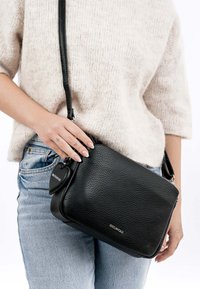 Persona, kas valkā gaišu džemperi un džinsus, turot melnu teksturētu šķērsgriezuma somiņu ar sirds formas atslēgu piekariņu un zīmola logotipa detaļu.