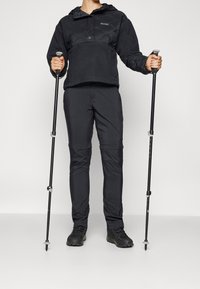 Sudadera negra aislante con capucha, forro polar en varias capas y diseño texturizado. Pantalones negros con bolsillos laterales y secciones ajustables, acompañados de bastones de senderismo.