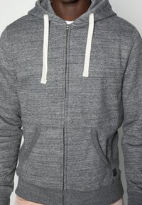 Grijze zip-up hoodie met witte trekkoorden, voorzakken en een gestructureerde stof. Heeft ribbelmanchetten en een klein zwart accentlogo.