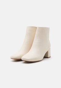 Bottines blanches en similicuir avec bout carré, talon bloc épais et design sans coutures. Texture lisse avec une silhouette minimaliste.