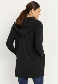Schwarze, mit Kapuze versehene, langärmlige Strickjacke aus strukturiertem Stoff mit Rippendesign und seitlichen Taschen, getragen über Jeanshose, von hinten betrachtet.