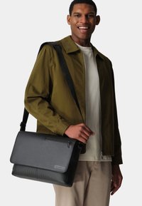 Schwarze strukturierte Umhängetasche mit einem Reißverschlussfach und verstellbarem Tragegurt, gehalten von einer Person in einer grünen Jacke und beigen Hosen.