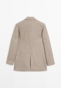 Cappotto beige su misura con maniche lunghe, colletto a revers, spacco posteriore e tessuto strutturato. Linee pulite, senza chiusure o motivi visibili.