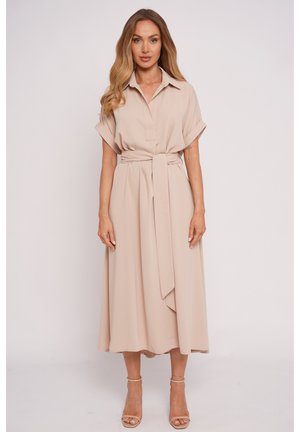 Kobieta ubrana w beżową, midi sukienkę z krótkim rękawem i kołnierzykiem, związaną w talii paskiem, stojąca z rękami swobodnie ułożonymi wzdłuż ciała, ubrana w beżowe szpilki.
