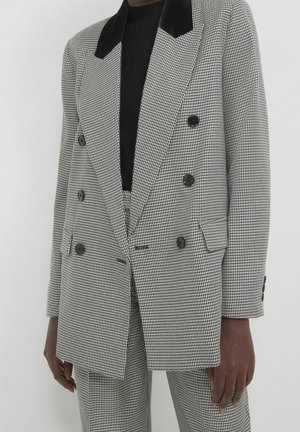 Person trägt einen schwarz-weißen Doppelreiher-Blazer im Hahnentrittmuster mit passenden Hosen und einem schwarzen Oberteil darunter, steht vor einem weißen Hintergrund.