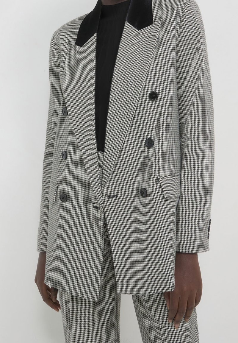 Personne portant un blazer croisé pied-de-poule noir et blanc avec un pantalon assorti et un haut noir en dessous, debout devant un fond blanc.