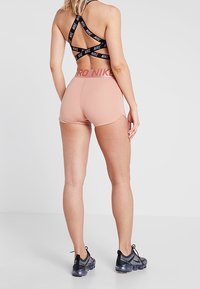 Žena v černém sportovním podprsenkovém topu Nike s křížovým střihem, růžových sportovních šortkách Pro Nike a šedých teniskách Nike, stojící zády k divákovi.