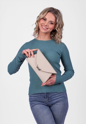 Beige Clutch-Tasche aus strukturiertem Material, mit einem dreieckigen Klappverschluss und einem dekorativen metallischen Akzent. Modell hält die Tasche.