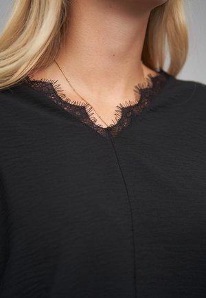 Schwarzes Oberteil aus strukturiertem Stoff, V-Ausschnitt mit Spitzenbesatz und feinen Fransen, sowie eine dezente Naht in der Mitte. Schlichte goldene Halskette sichtbar.