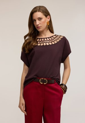 Vrouw met lang bruin haar die een donkerpaarse top met een ronde uitsnede bij de hals draagt, rode broek met zwarte riem en grote oorbellen.