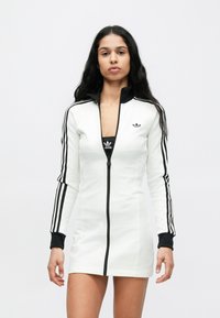 Λευκό αθλητικό φόρεμα με μαύρο φερμουάρ και γιακά, με τρεις κάθετες μαύρες ρίγες σε κάθε μανίκι και ένα μικρό λογότυπο στο στήθος.
