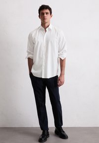 Witte button-up shirt met opgerolde mouwen, gecombineerd met marineblauwe broeken en zwarte veterschoenen. Gladde stof, klassieke kraag en knoopdetails.