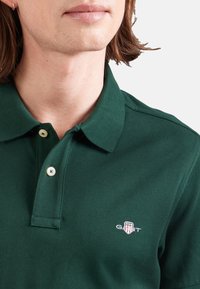 Ciemnozielona koszulka polo o teksturowanym wykończeniu, z dwoma guzikami i małym haftowanym logo na piersi.