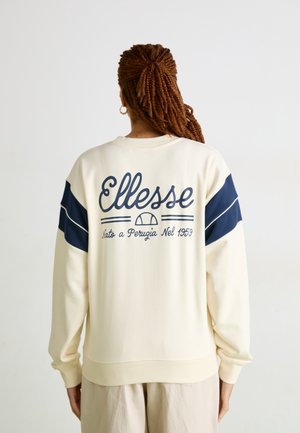 Personne aux cheveux tressés porte un sweat crème avec des rayures bleu marine et le logo Elle sse ainsi qu’un texte au dos sur un fond uni.