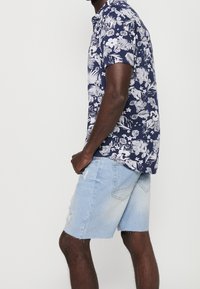 Homme portant une chemise à manches courtes bleu marine à motif floral et un short en jean bleu clair, debout de profil contre un fond clair uni.