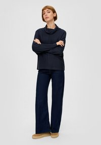 Navy cowl-neck top in ribbelstof, gecombineerd met wijde, donkere spijkerbroeken en tan platform sneakers, wat zorgt voor een ontspannen silhouet.