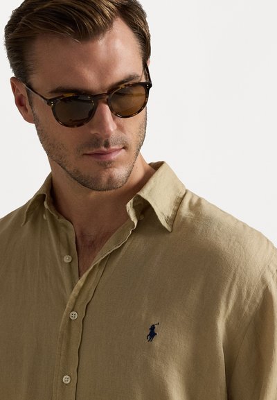 Camisa de linho bege com botões, apresentando uma textura subtil e um pequeno logótipo escuro no lado esquerdo do peito. O modelo usa óculos de sol castanhos.