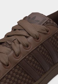 Καφέ πλεκτό δερμάτινο αθλητικό παπούτσι με χοντρές κορδόνια, τρεις χαρακτηριστικές ρίγες και το λογότυπο τριφύλλι της Adidas στη γλώσσα.