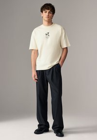 Cream shirt with short sleeves and black floral graphic. Loose-fitting black trousers and black shoes complete the outfit. Simple backdrop.