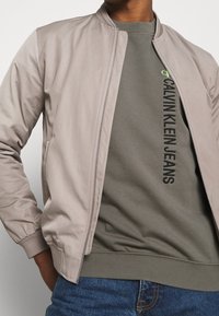 Chaqueta bomber beige claro de textura suave, con cierre de cremallera y puños acanalados, llevada sobre una sudadera verde con texto negro de marca.