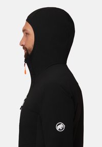 Black softshell jacket with a hood, featuring a zippered chest pocket and a white logo on the left sleeve. Smooth, matte texture.
