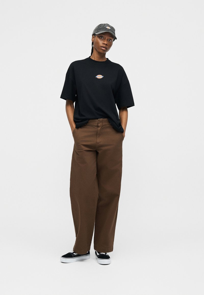 Donna che indossa una maglietta nera con logo, pantaloni marroni a gamba larga, berretto nero e scarpe da ginnastica nere, in piedi con le mani nelle tasche davanti a uno sfondo semplice.