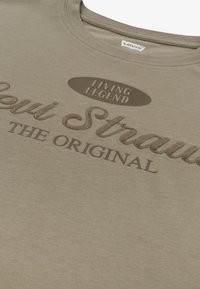 Χακί μπλουζάκι με καφέ ανάγλυφη γραφή. Το σχέδιο περιλαμβάνει τις φράσεις "Living Legend" και "Levi Strauss THE ORIGINAL" σε διαφορετικές γραμματοσειρές. Κυκλικό λογότυπο.