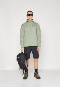 Šviesiai žalia zip-up striukė, juodas kuprinės, juodos šortai, žygių batai. Yra gobtuvas, logotipas ant krūtinės ir kontrastingos tekstūros.