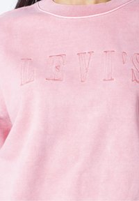 Sweat-shirt rose avec le logo "LEVI'S" brodé en relief sur la poitrine, porté par une personne avec un cou visible et des mèches de cheveux.